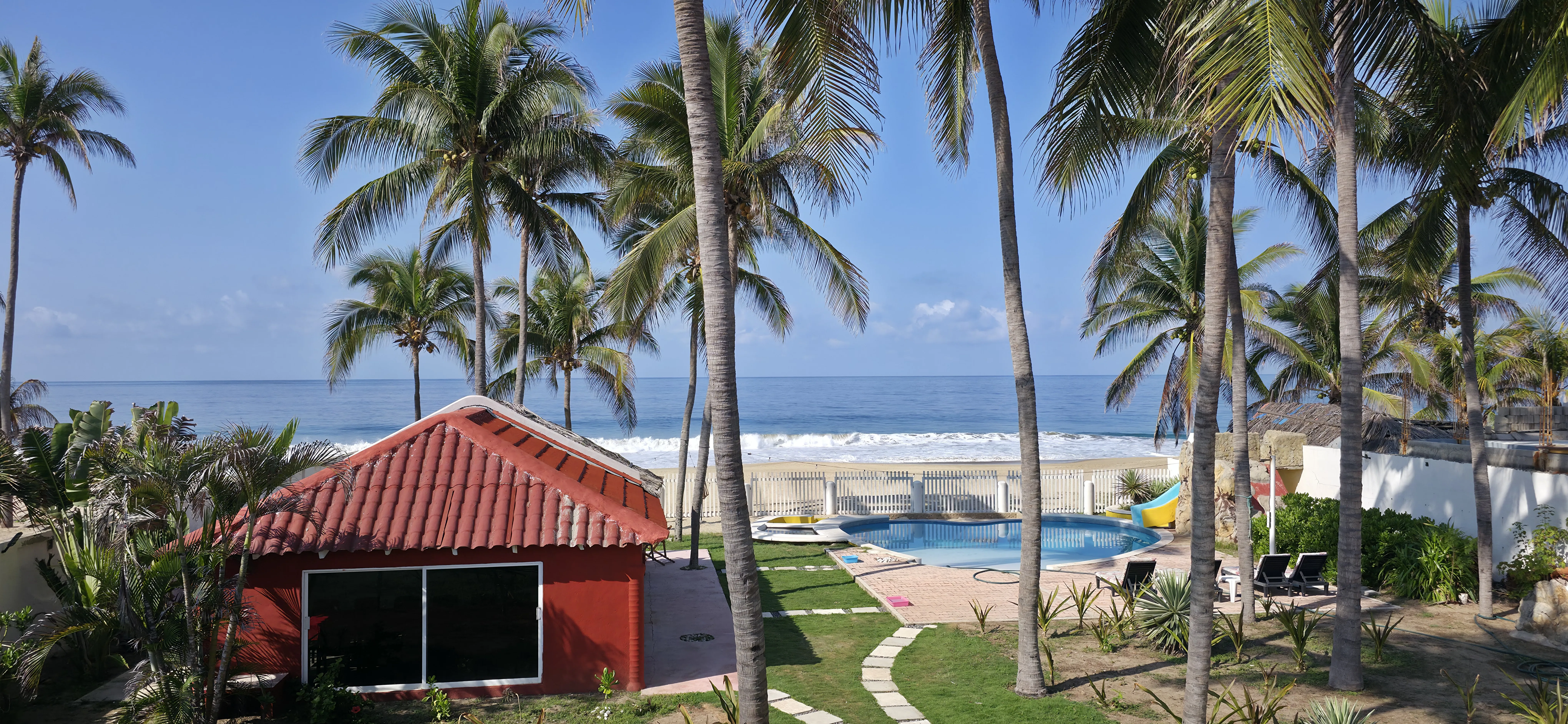 Vista elevada de la alberca con palapa de tejas rojas y el océano al fondo